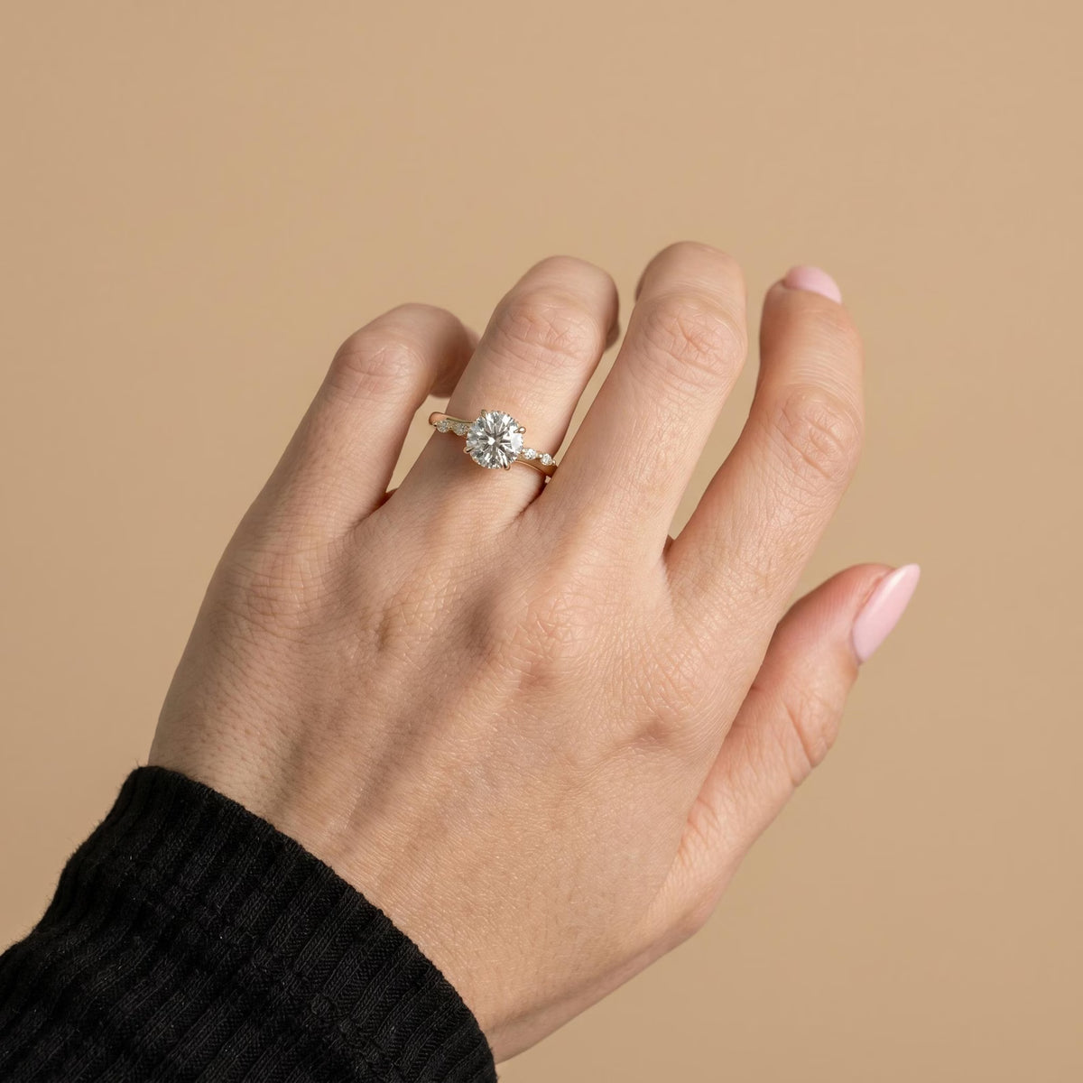 A close-up of a hand wearing a delicate gold ring with a small clear gemstone cluster, set against a solid beige background.