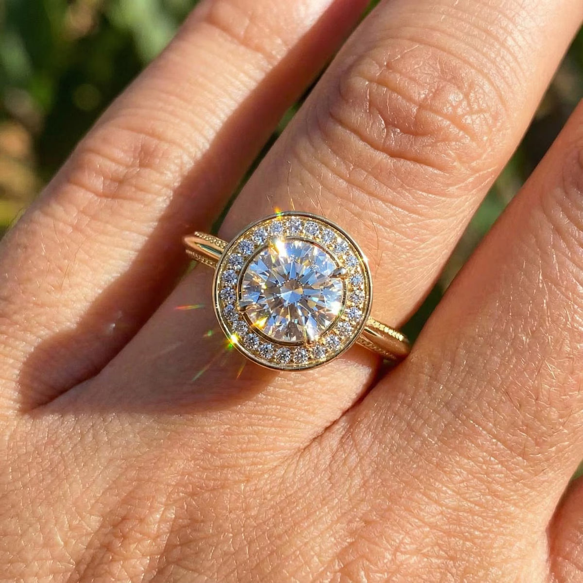 A gold ring with a round brilliant-cut center stone and a halo of small diamonds, worn on a person's finger outdoors with blurred green foliage in the background.