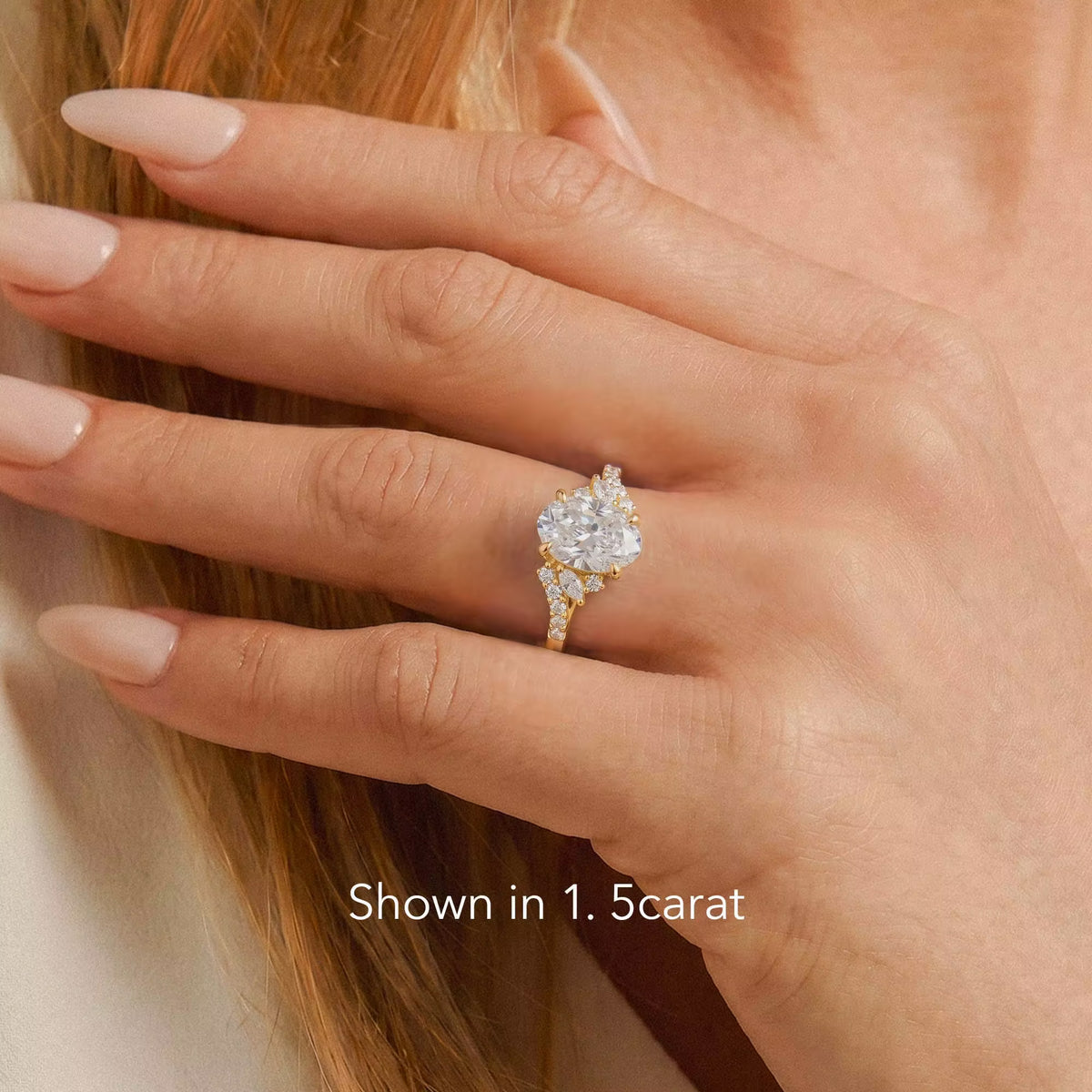 A close-up of a hand wearing a yellow gold ring with a large round-cut clear gemstone and smaller accent stones. The hand is resting against light skin and light brown hair, with the text 'Shown in 1.