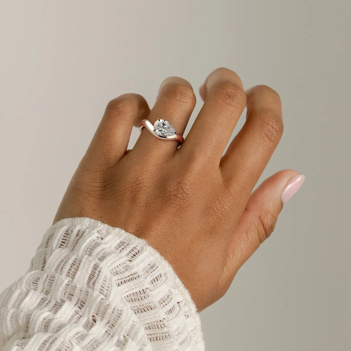 A close-up of a hand wearing a silver ring with a small clear gemstone, styled with a white textured knit sweater against a neutral light gray background.